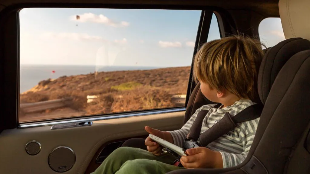 Young child in a car seat looking out a tinted rear side window at a sunny coastline