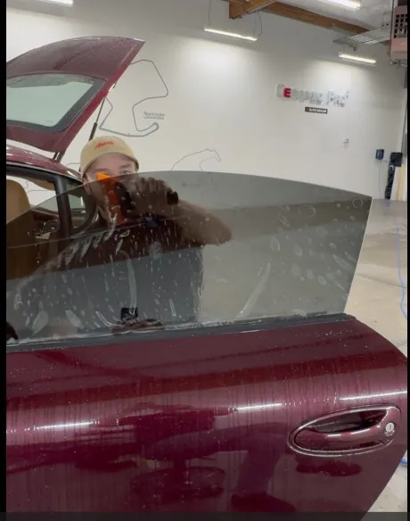 APC Auto Spa installer in the Tukwila shop applying window tint film to the wet inside of a maroon car's door window